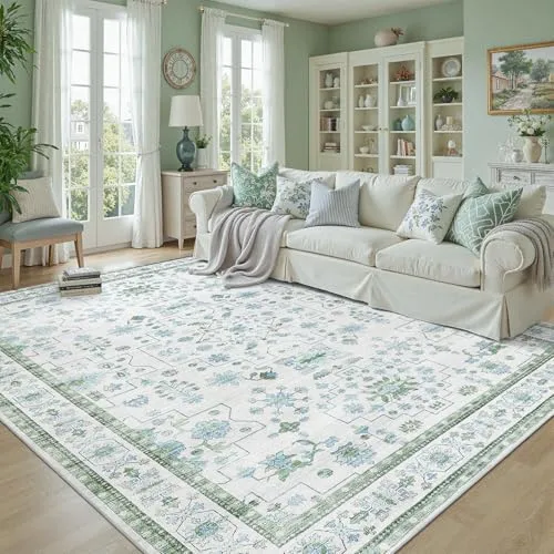 A living room features a light-colored sofa with green and floral accent pillows, a textured throw blanket, a large rectangular area rug with a floral pattern, a side table with books, and a lamp, all against a backdrop of large windows and a display cabinet.
