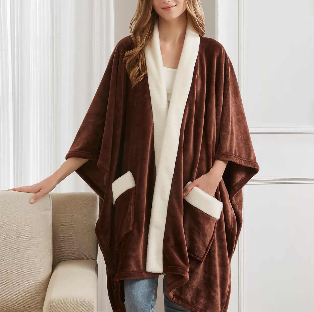 A woman is wearing a brown, fleece-lined, open-front poncho with white trim and pockets, standing beside a beige couch.