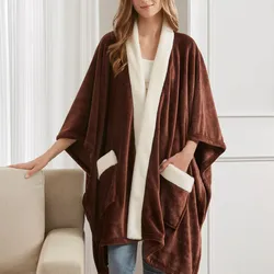 A woman is wearing a brown, fleece-lined, open-front poncho with white trim and pockets, standing beside a beige couch.