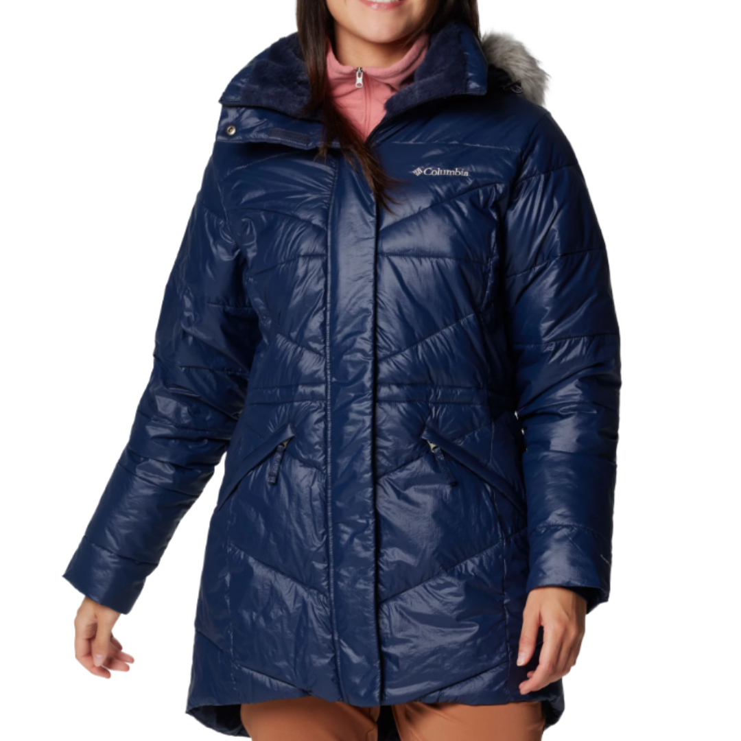 A woman is wearing a blue Columbia puffer coat with a fur-lined hood and zippered pockets, paired with a pink turtleneck and brown pants.