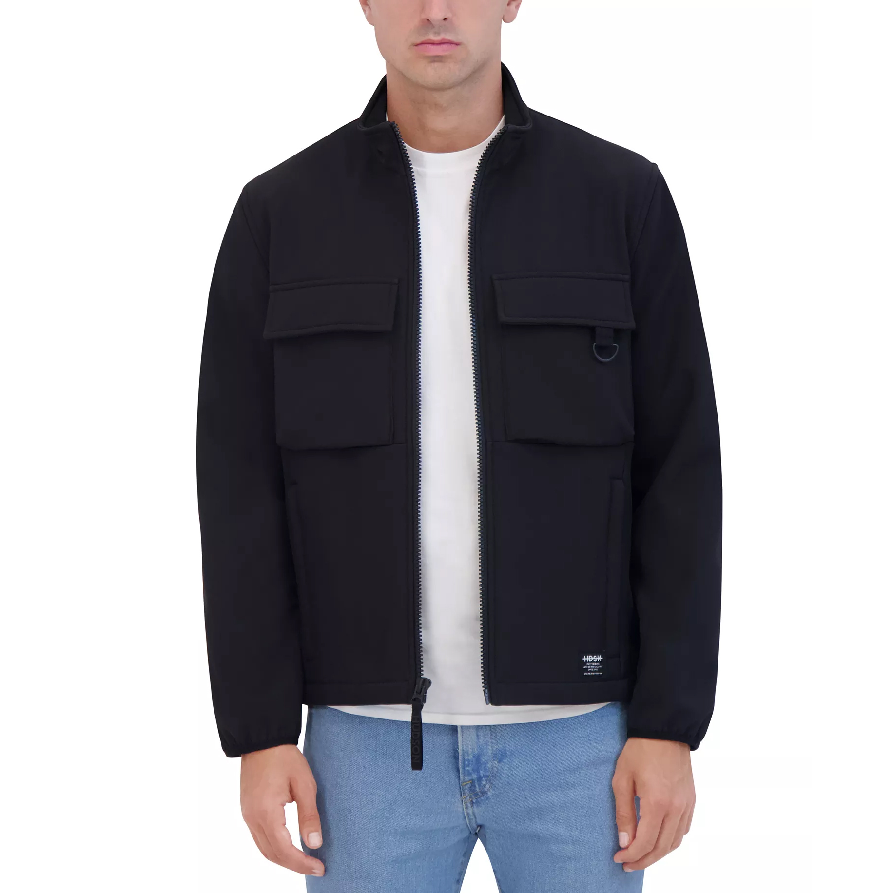 A man is wearing a black utility jacket with front pockets and a zipper closure, paired with a white t-shirt and light blue jeans.