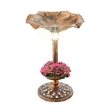 A bronze-colored birdbath with a solar light on top, featuring a design resembling a leaf. It includes a lower section filled with pink flowers and a decorative base.