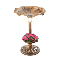 A bronze-colored birdbath with a solar light on top, featuring a design resembling a leaf. It includes a lower section filled with pink flowers and a decorative base.