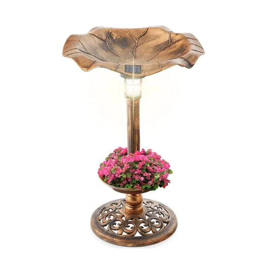 A bronze-colored birdbath with a solar light on top, featuring a design resembling a leaf. It includes a lower section filled with pink flowers and a decorative base.
