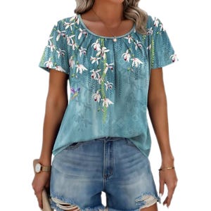 A light teal short-sleeve boho top with a floral pattern of white flowers and greenery cascading from the neckline. The top is paired with distressed denim shorts.