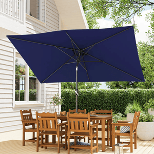 Patio set with a rectangular wooden table and six chairs under a large navy blue rectangular umbrella, situated on a deck beside a white house with greenery in the background.
