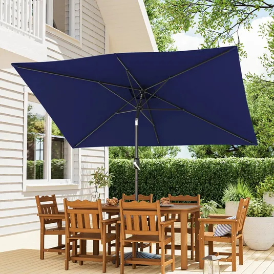 Patio set with a rectangular wooden table and six chairs under a large navy blue rectangular umbrella, situated on a deck beside a white house with greenery in the background.
