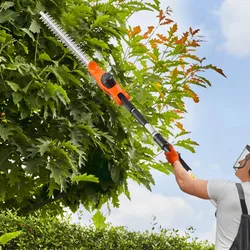 A person is using an orange and black telescopic hedge trimmer to cut tall green foliage while wearing protective gear, including a face shield and gloves.