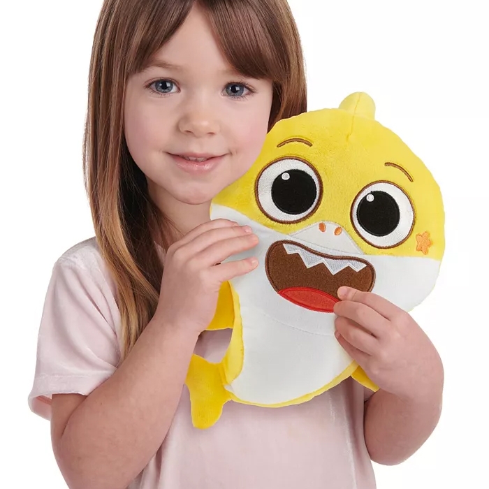 A young girl is holding a yellow and white shark plush toy with expressive eyes and an open mouth.