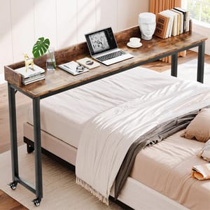 An overbed table with metal legs and a rustic wooden top is positioned above a bed. It's equipped with wheels and holds items like a laptop, plants, books, and a mug.