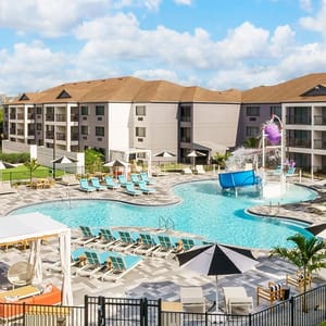 A resort pool area features a swimming pool, lounge chairs, umbrellas, a splash pad with a waterslide, and nearby apartment buildings under a blue sky with clouds.