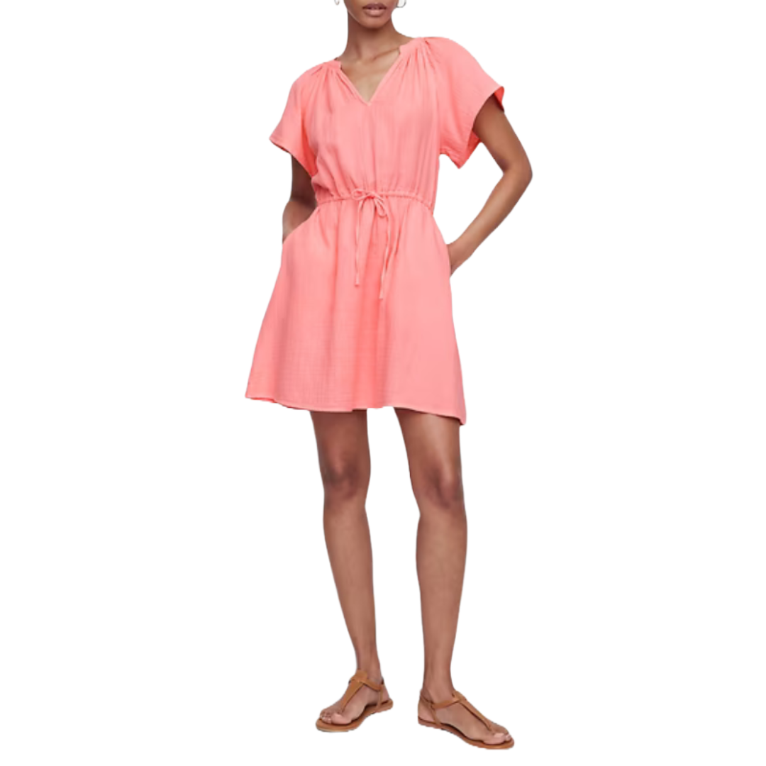 Coral pink gauze splitneck dress with a V-neckline, short sleeves, and a cinched waist tie. It's knee-length and paired with flat sandals.