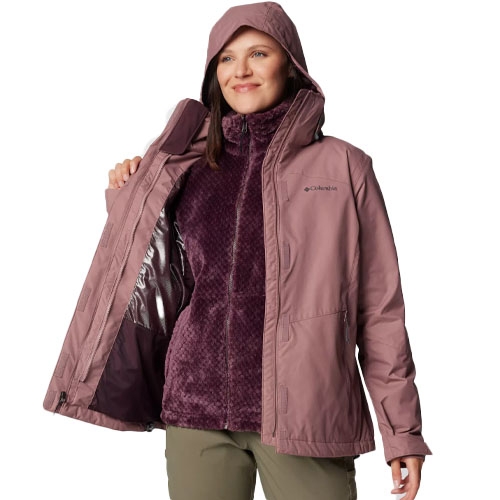 A woman wears a two-layer winter jacket with a hood, featuring an outer shell and an inner plush, textured lining. The jacket is mauve, and she is dressed in olive green pants.