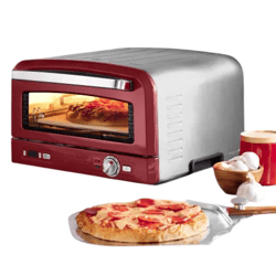 A red and silver countertop pizza oven is shown next to a cooked pizza, garlic, and spices on a kitchen counter.