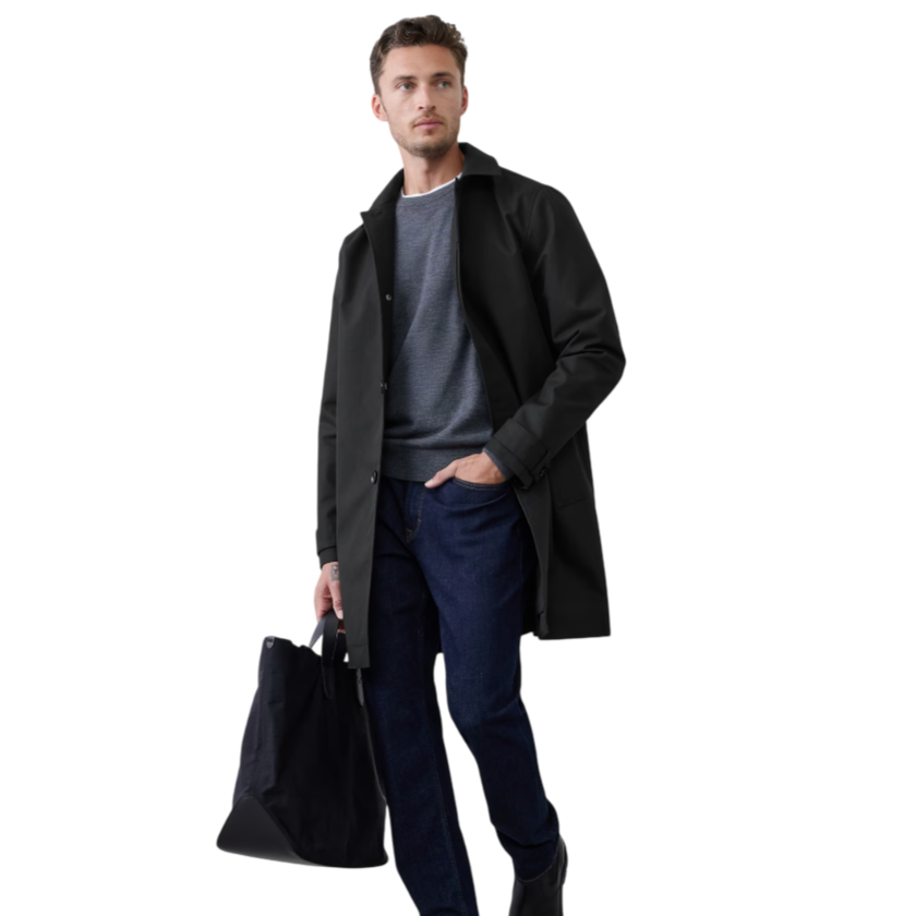 A man wears a black coat over a gray sweater with dark blue jeans and black boots, holding a black tote bag.