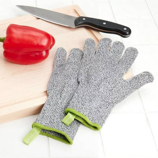 A pair of cut-resistant gloves with a gray knit construction and green cuffs is laid out beside a kitchen knife and red bell pepper on a wooden cutting board.