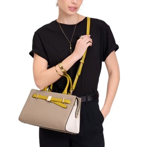 A taupe and white leather satchel with yellow accents, featuring dual handles, a detachable shoulder strap, and a front decorative buckle detail. Worn by a model in a black outfit with gold accessories.