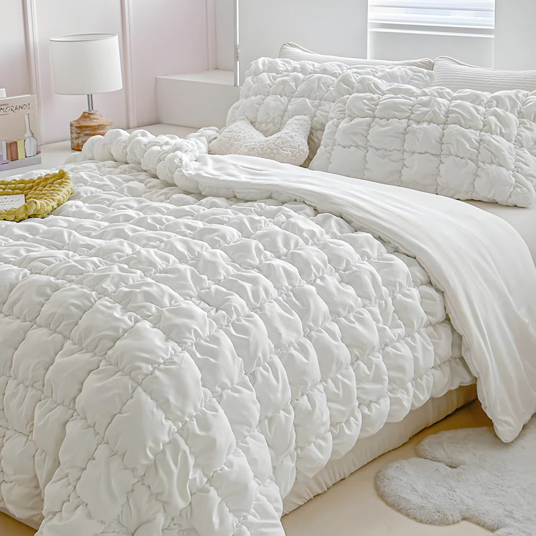 A neatly made bed features a quilted white comforter, matching pillows, and a soft heart-shaped cushion. A small side table holds a lamp, and a plush rug rests on the floor.