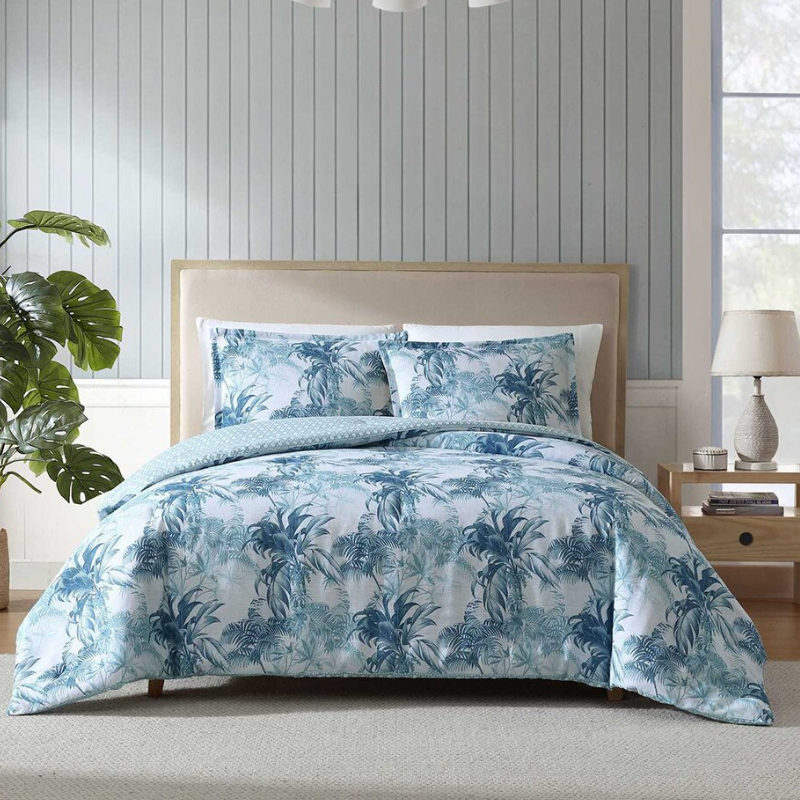 A bed setup with a tropical-themed blue and white duvet cover and matching pillowcases. The room features a wooden nightstand with a lamp and plant accents.