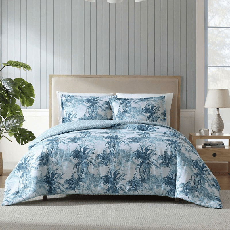 A bed setup with a tropical-themed blue and white duvet cover and matching pillowcases. The room features a wooden nightstand with a lamp and plant accents.