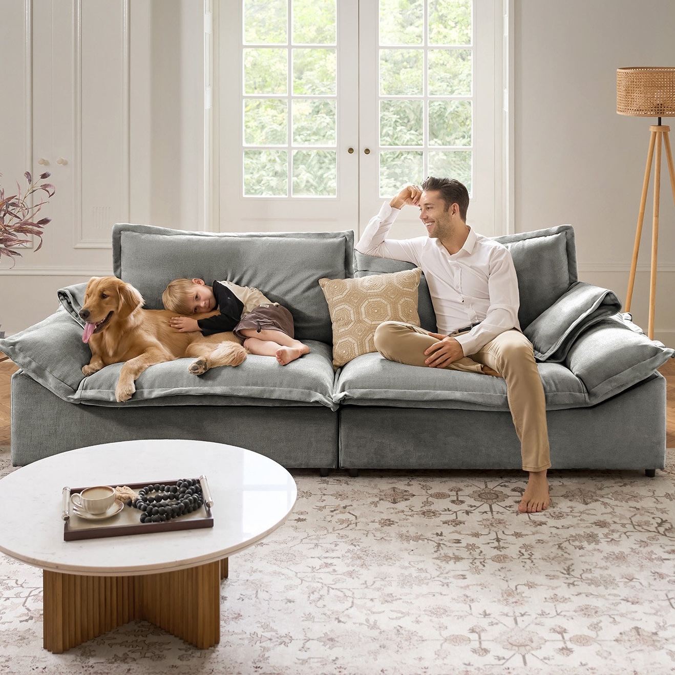 A man, child, and dog are on a plush, gray sofa in a cozy living room. The sofa is against a bright window, with a round coffee table and decorative items nearby.