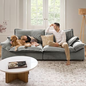 A man, child, and dog are on a plush, gray sofa in a cozy living room. The sofa is against a bright window, with a round coffee table and decorative items nearby.