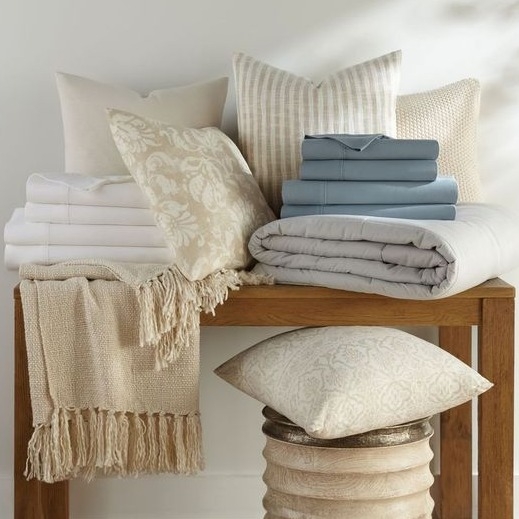A collection of bedding items including beige and white pillows, folded blue and white sheets, a cozy knit throw with fringe, and a quilt, all arranged on and around a wooden bench.