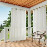Outdoor patio curtains hang from a wooden ceiling on a porch, offering privacy and shade. A wicker chair and a small side table complete the setting.