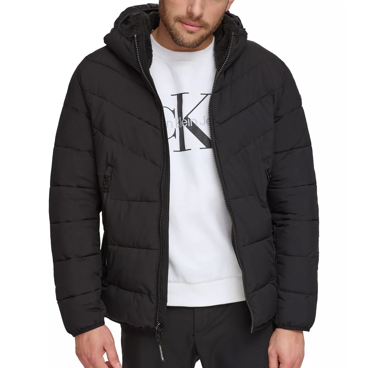 A man is wearing a black puffer jacket with a hood and a white sweatshirt featuring the Calvin Klein logo.
