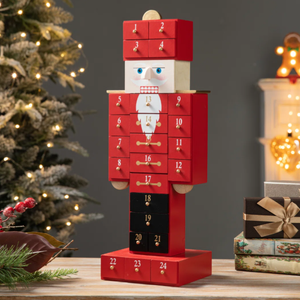 A wooden nutcracker-themed advent calendar with numbered drawers for each day leading up to Christmas, surrounded by festive decorations including a lit Christmas tree and wrapped gifts.