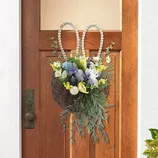 A wicker basket decorated with a floral arrangement, featuring purple, white, and yellow flowers, and greenery, is hanging on a door. It has beaded loops resembling bunny ears at the top.