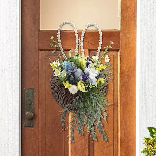 A wicker basket decorated with a floral arrangement, featuring purple, white, and yellow flowers, and greenery, is hanging on a door. It has beaded loops resembling bunny ears at the top.