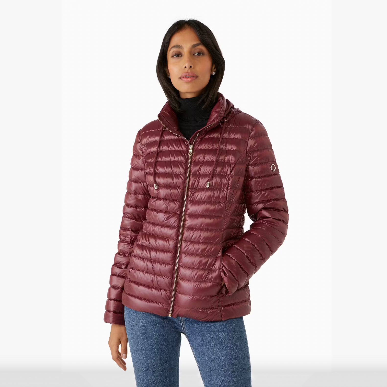 A woman is wearing a burgundy down jacket with a high collar and horizontal quilting, enhanced by a subtle sheen. The jacket has a zip closure and a logo on the left sleeve.