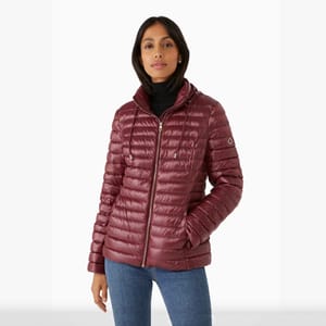 A woman is wearing a burgundy down jacket with a high collar and horizontal quilting, enhanced by a subtle sheen. The jacket has a zip closure and a logo on the left sleeve.