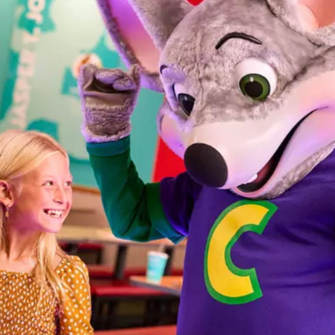 A child smiles next to a person in a mouse costume with a purple shirt featuring a yellow \“C\“. They are in an indoor play area with a colorful background.