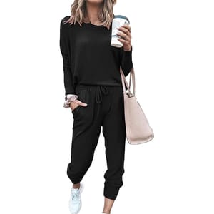 A woman is wearing a black long-sleeve jumpsuit with drawstring waist, holding a coffee cup, and carrying a light pink handbag. She has white sneakers and a pink scrunchie on her wrist.