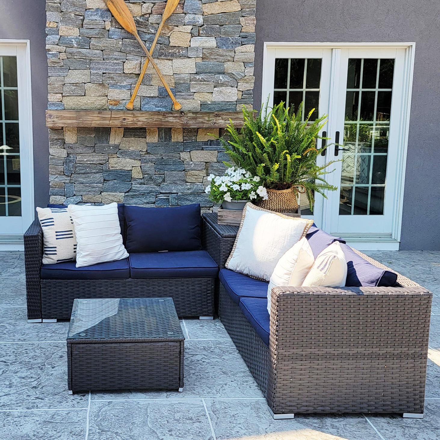 Outdoor patio set with navy cushions, wicker sectional sofa, and a glass-top coffee table on a stone floor. Background features a stone wall with crossed wooden oars and a shelf with plants.