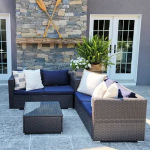 Outdoor patio set with navy cushions, wicker sectional sofa, and a glass-top coffee table on a stone floor. Background features a stone wall with crossed wooden oars and a shelf with plants.