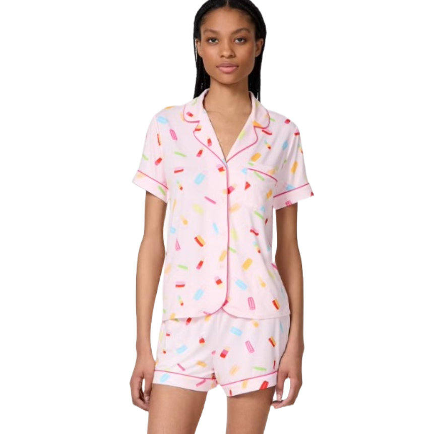A woman is wearing a pink pajama set with a playful multicolored popsicle print, featuring a short-sleeve button-up shirt and matching shorts.