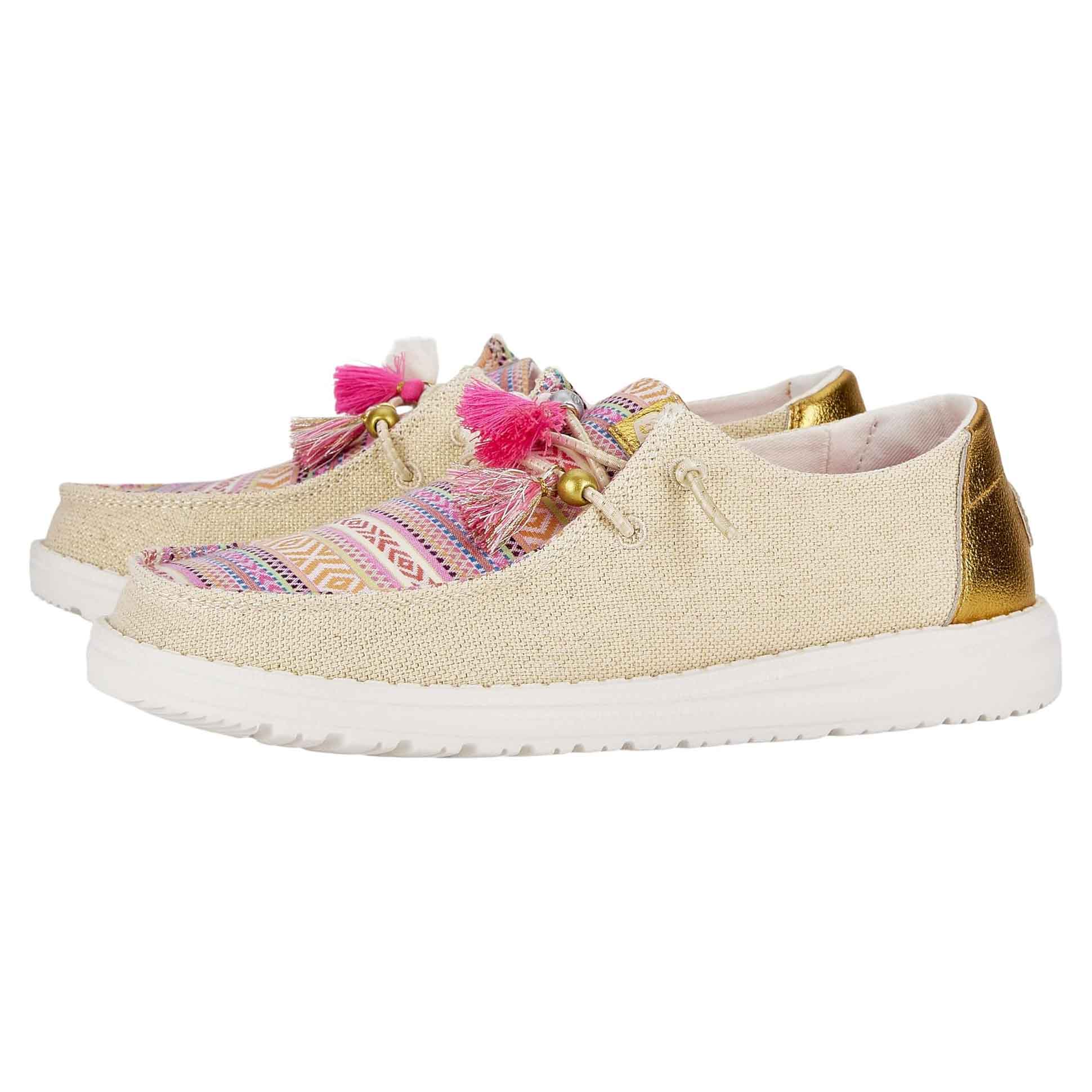 Hey Dude Wendy Boho Festival Shoes feature a beige fabric with colorful woven patterns, bright pink tassels, and gold accents on the heel. They have white soles and a casual, slip-on design.