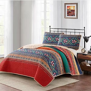 A colorful quilt set with matching pillows on a bed, situated in a room with curtains, artwork, and a desk.