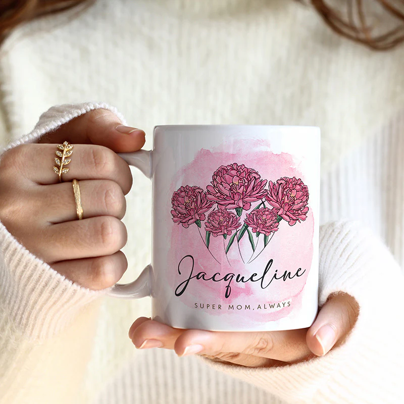 A person holds a white mug with pink flowers and the name \“Jacqueline\“ printed on it, along with the phrase \“Super Mom, Always.\“ The person wears a white sweater and gold rings.