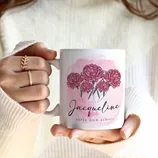 A person holds a white mug with pink flowers and the name \“Jacqueline\“ printed on it, along with the phrase \“Super Mom, Always.\“ The person wears a white sweater and gold rings.