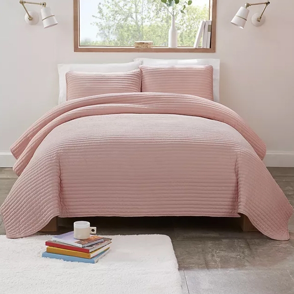 The UGG 3pc King Quilt Set features a blush-colored quilt with a striped texture, accompanied by two matching pillow shams. The bed is styled in a minimalist room with a window, white side lamps, and a small bookshelf on the floor.