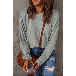 A woman is wearing a long-sleeved, relaxed-fit sage green top with a round neckline, paired with distressed blue jeans and a brown crossbody bag.