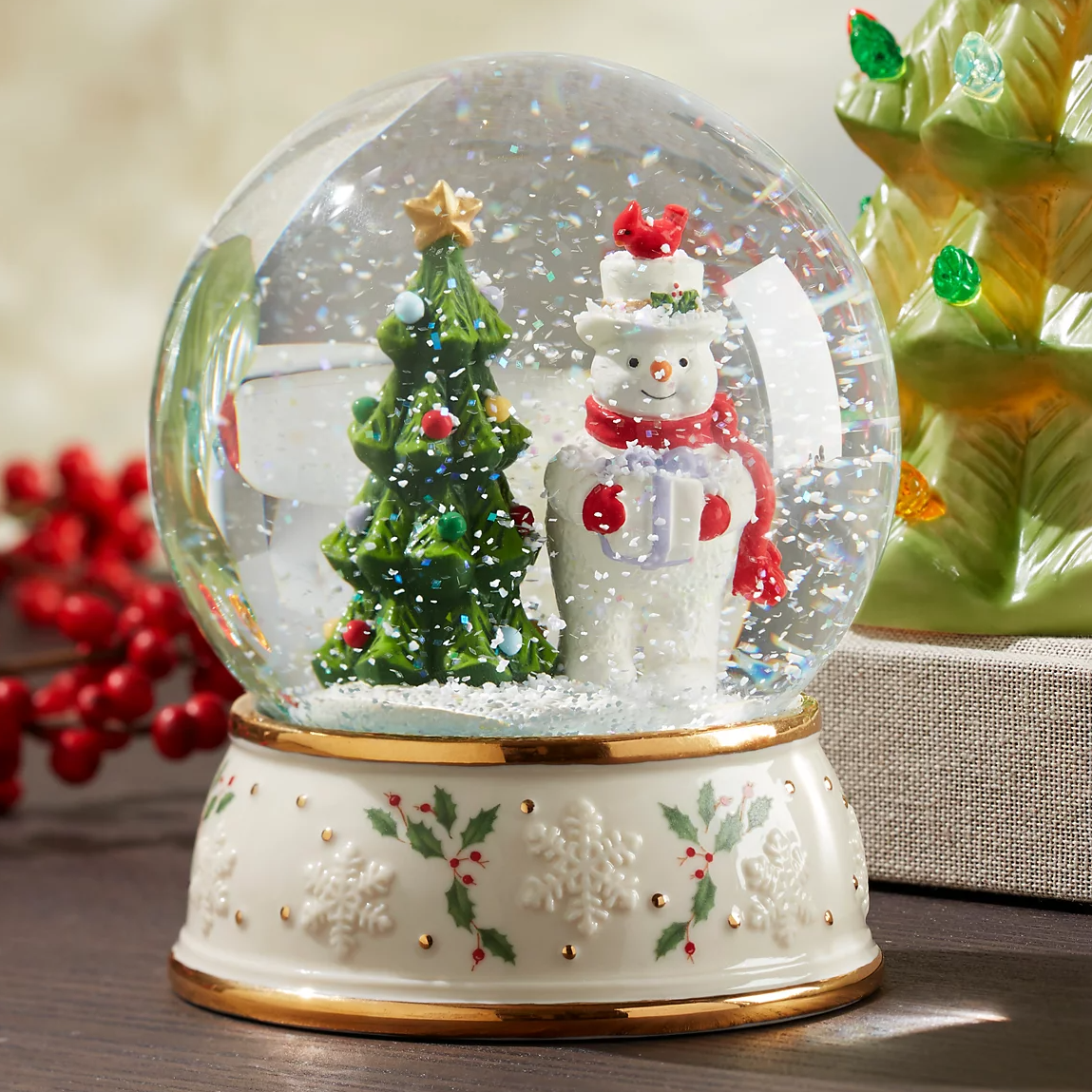 The Lenox Snowman Snow Globe features a festive scene with a snowman wearing a red scarf and a top hat next to a decorated Christmas tree. The base is adorned with holly and snowflake patterns.