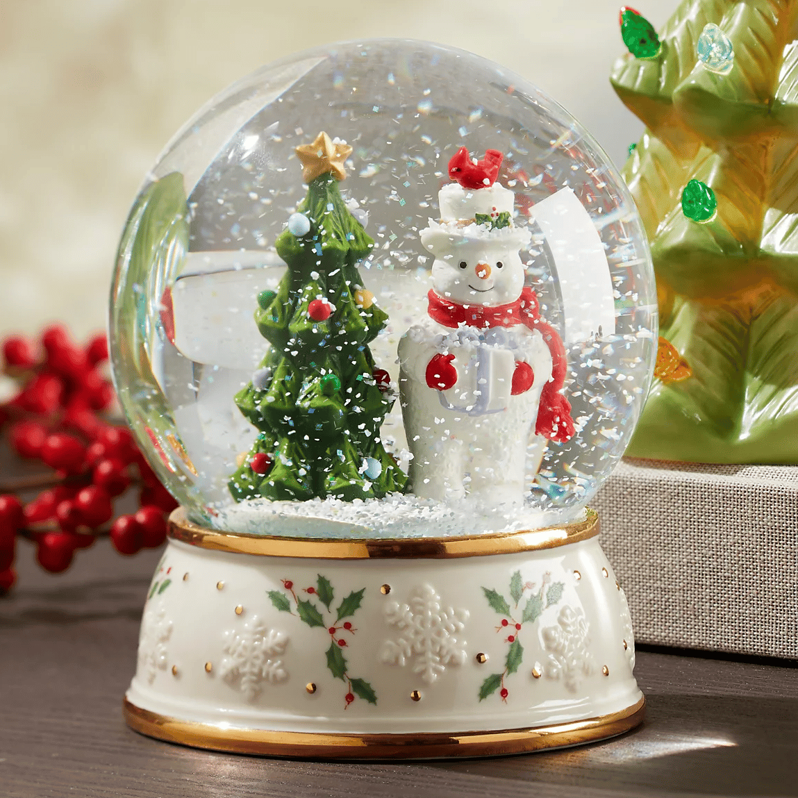 The Lenox Snowman Snow Globe features a festive scene with a snowman wearing a red scarf and a top hat next to a decorated Christmas tree. The base is adorned with holly and snowflake patterns.