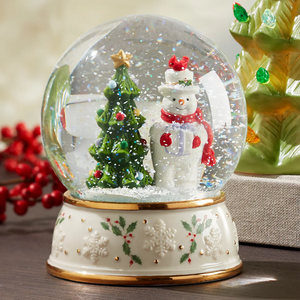 The Lenox Snowman Snow Globe features a festive scene with a snowman wearing a red scarf and a top hat next to a decorated Christmas tree. The base is adorned with holly and snowflake patterns.