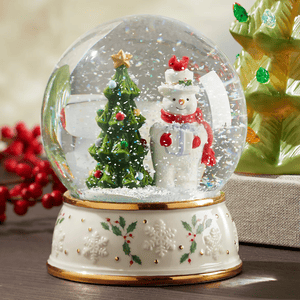 The Lenox Snowman Snow Globe features a festive scene with a snowman wearing a red scarf and a top hat next to a decorated Christmas tree. The base is adorned with holly and snowflake patterns.