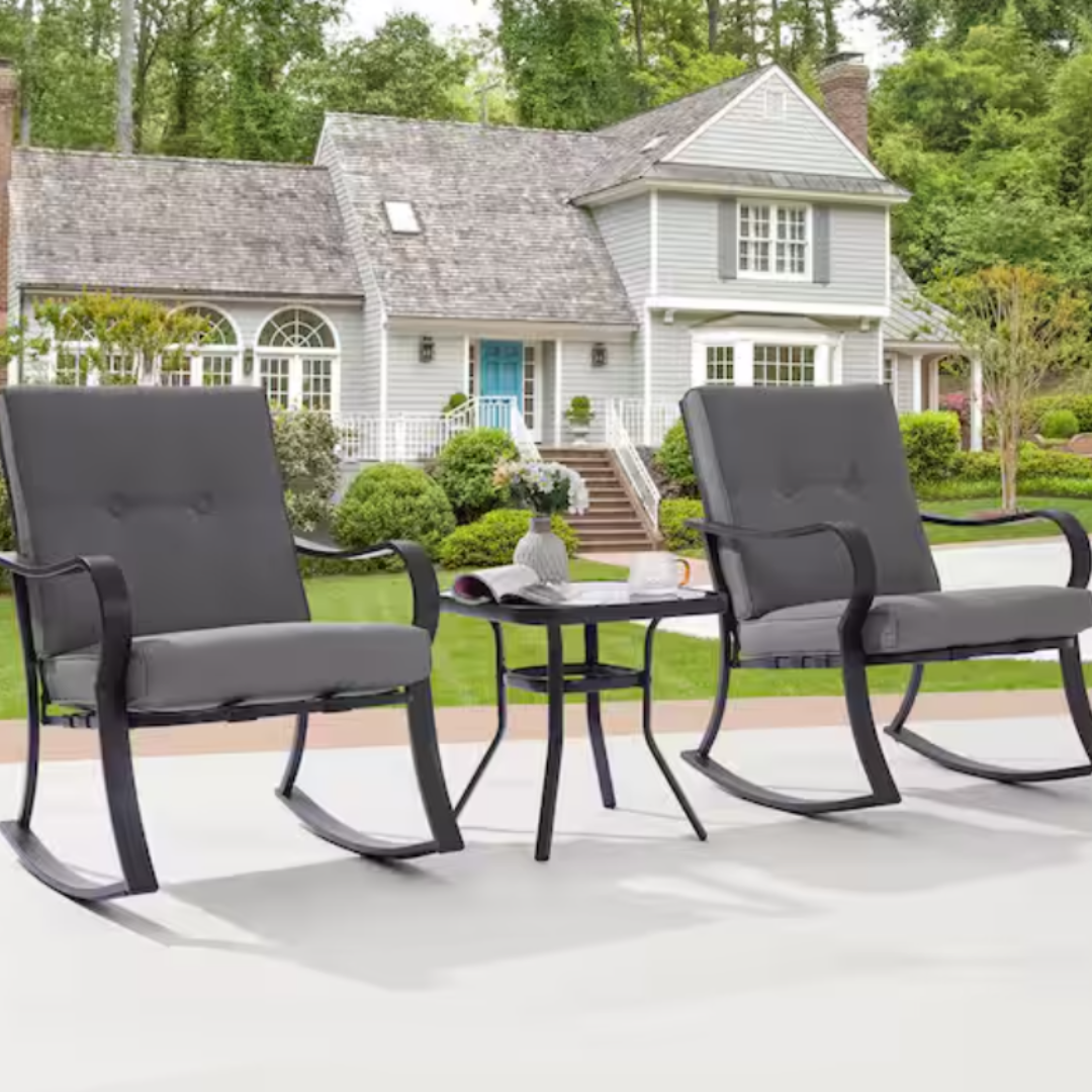 Two dark grey outdoor rocking chairs and a matching side table, placed on a patio with a house in the background.